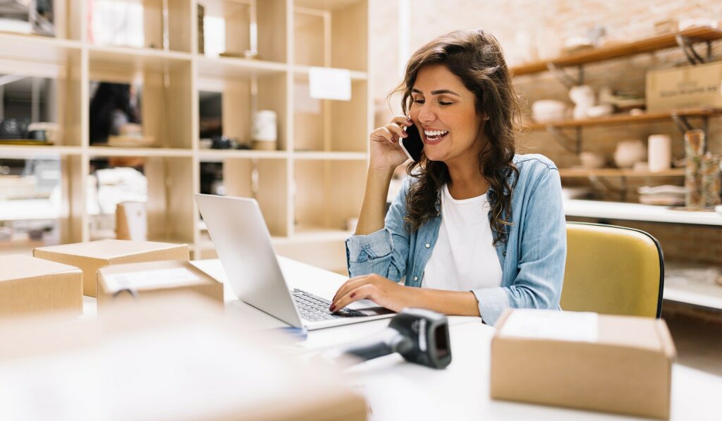 Mulher sentada falando no celular enquanto olha e mexe em um laptop