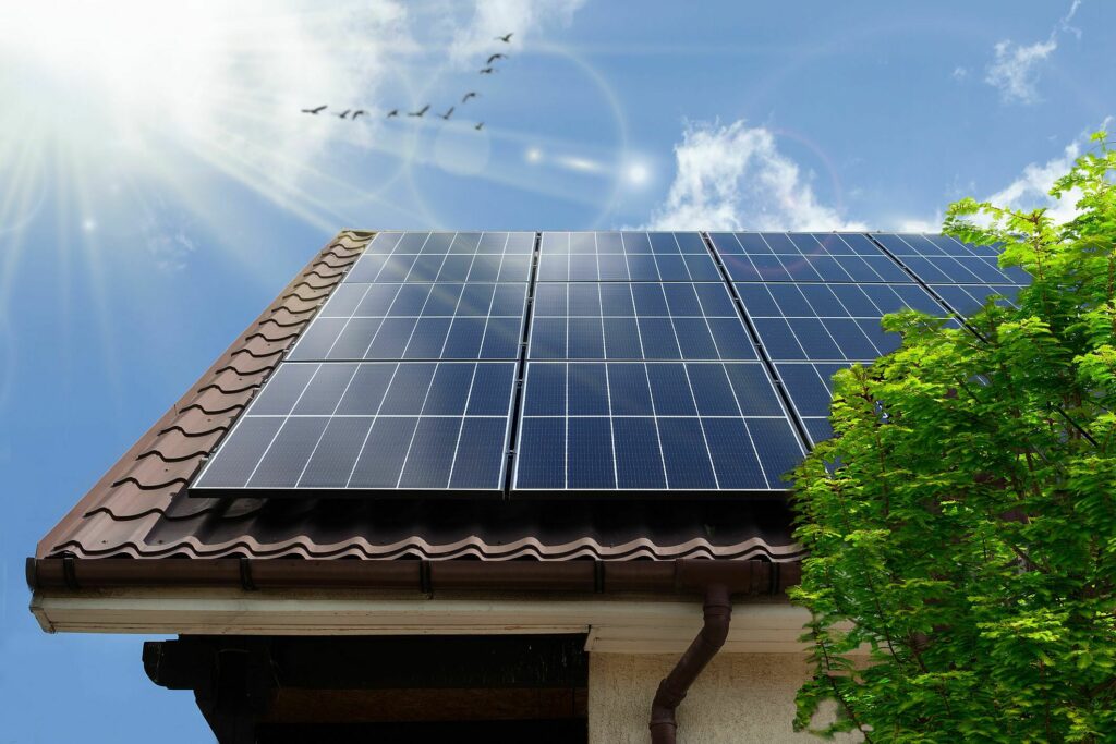 Telhado com painéis solares instalados e, ao fundo, o céu com poucas nuvens e o sol com efeito lens flare