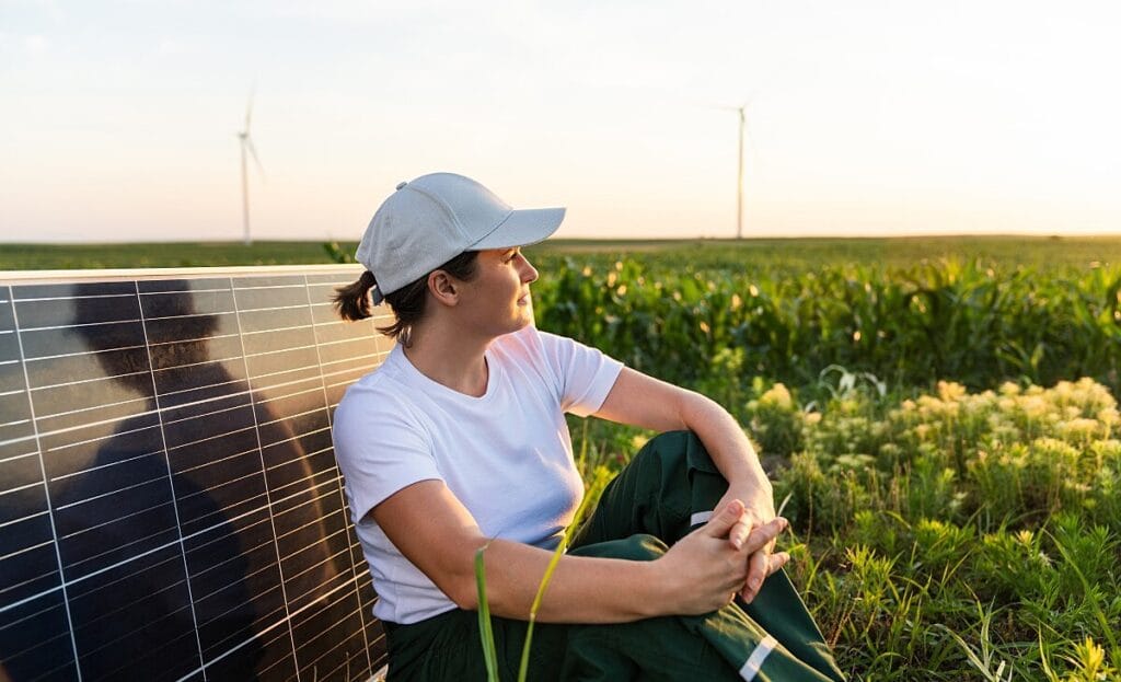 Mulher sentada no campo com plantação e um painel solar atrás dela